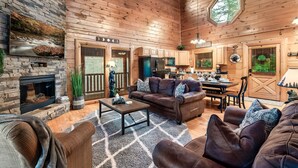 A cozy cabin interior with a stone fireplace, wooden walls, a dining area, and a living room furnished with leather sofas and a coffee table. - Mountain View/Hot Tubs/Pools/Game Room/Putt-Putt Course/Master Bedrooms (Sevierville)