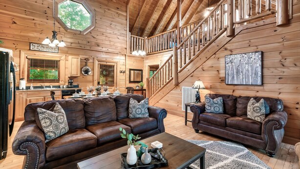 A spacious living area with two brown leather sofas, a wooden staircase, and a kitchen in the background. - Mountain View/Hot Tubs/Pools/Game Room/Putt-Putt Course/Master Bedrooms (Sevierville)