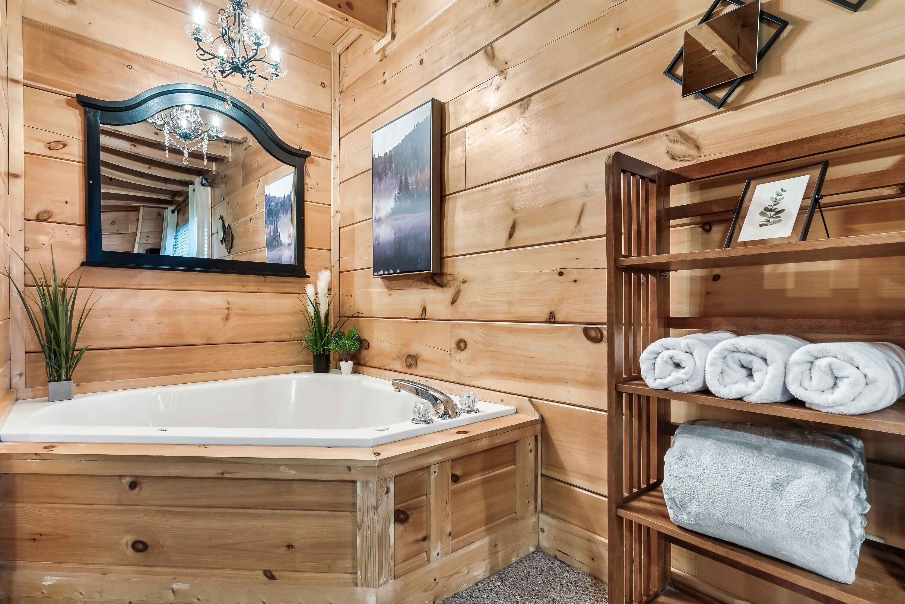 A wooden cabin bathroom with a large bathtub, a mirror, a chandelier, and shelves with towels.