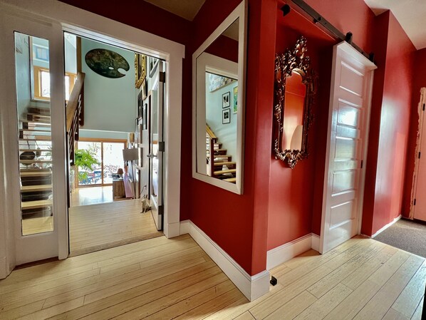 Un couloir avec un plancher en bois, un mur rouge et une porte blanche. Un miroir est accroché au mur, et un escalier mène à une autre pièce.