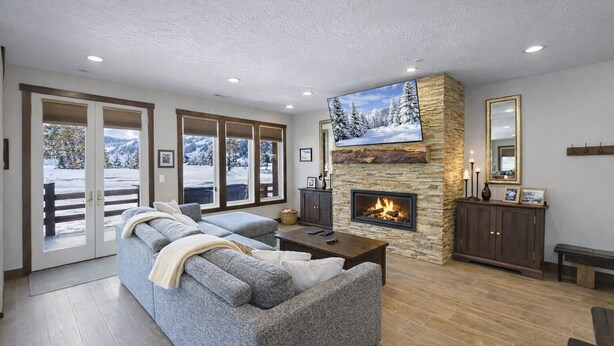 Una sala de estar acogedora con chimenea de piedra, un televisor de pantalla plana grande y vistas a un paisaje nevado a través de puertas de vidrio.