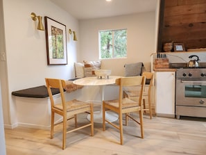 A cozy kitchen with a round table, wooden chairs, a stove, and a window with a view of trees.