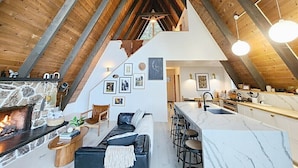 Een ruime loft met hoge plafonds, zichtbare houten balken, een stenen openhaard, een moderne keuken met een marmeren aanrechtblad en een gezellig zithoek met een bank en stoelen.