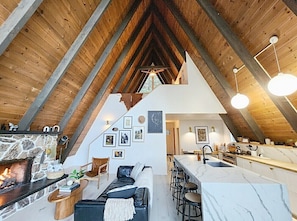 A spacious loft with a high ceiling, exposed wooden beams, a stone fireplace, a modern kitchen with a marble countertop, and a cozy seating area with a sofa and chairs.