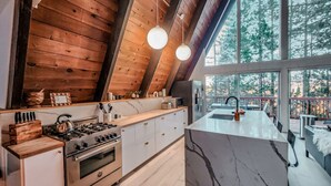 Een moderne keuken met een groot eiland, houten balken in het plafond en uitzicht op bomen door grote ramen.