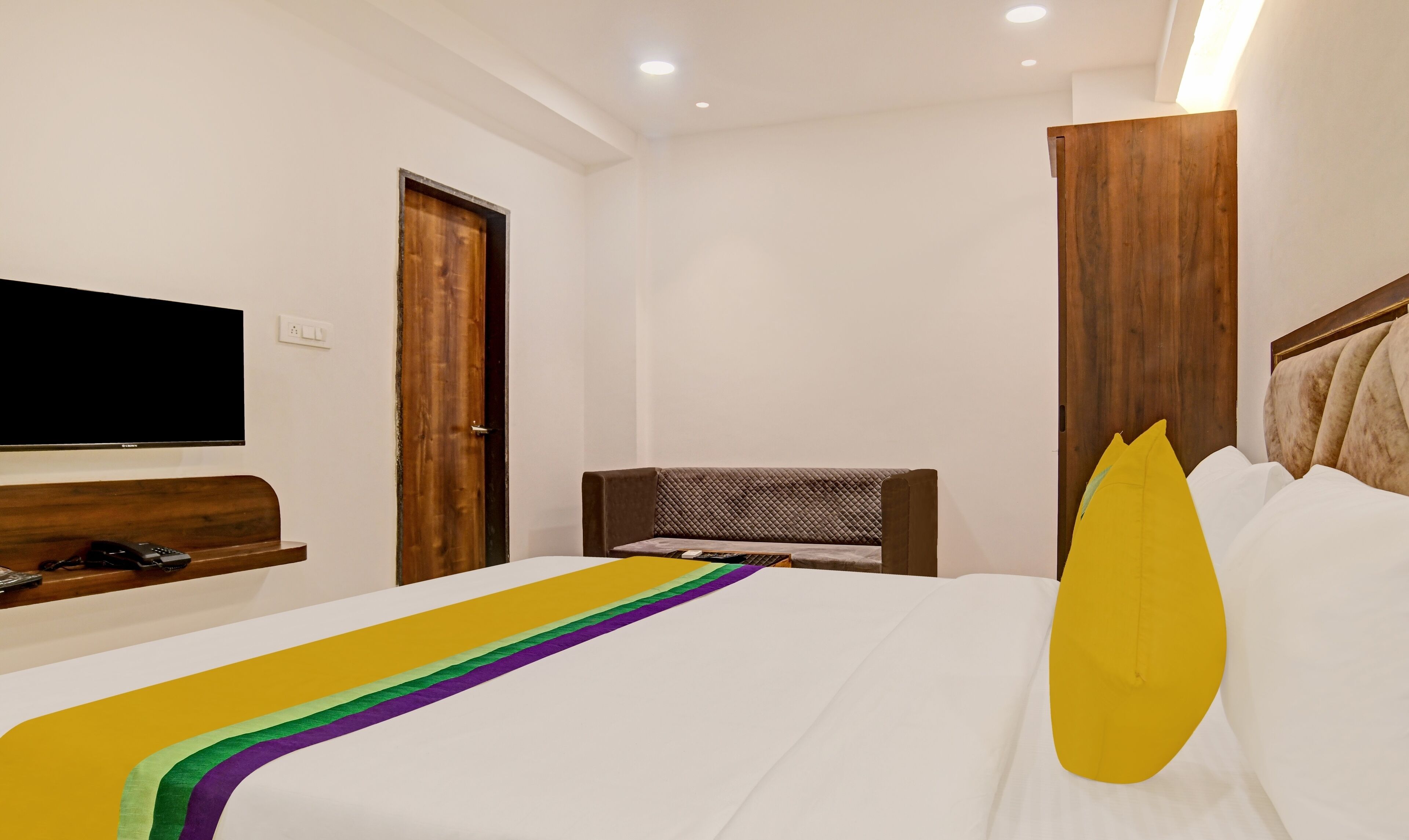 A hotel room with a bed, a TV mounted on the wall, a wooden door, and a wooden headboard.