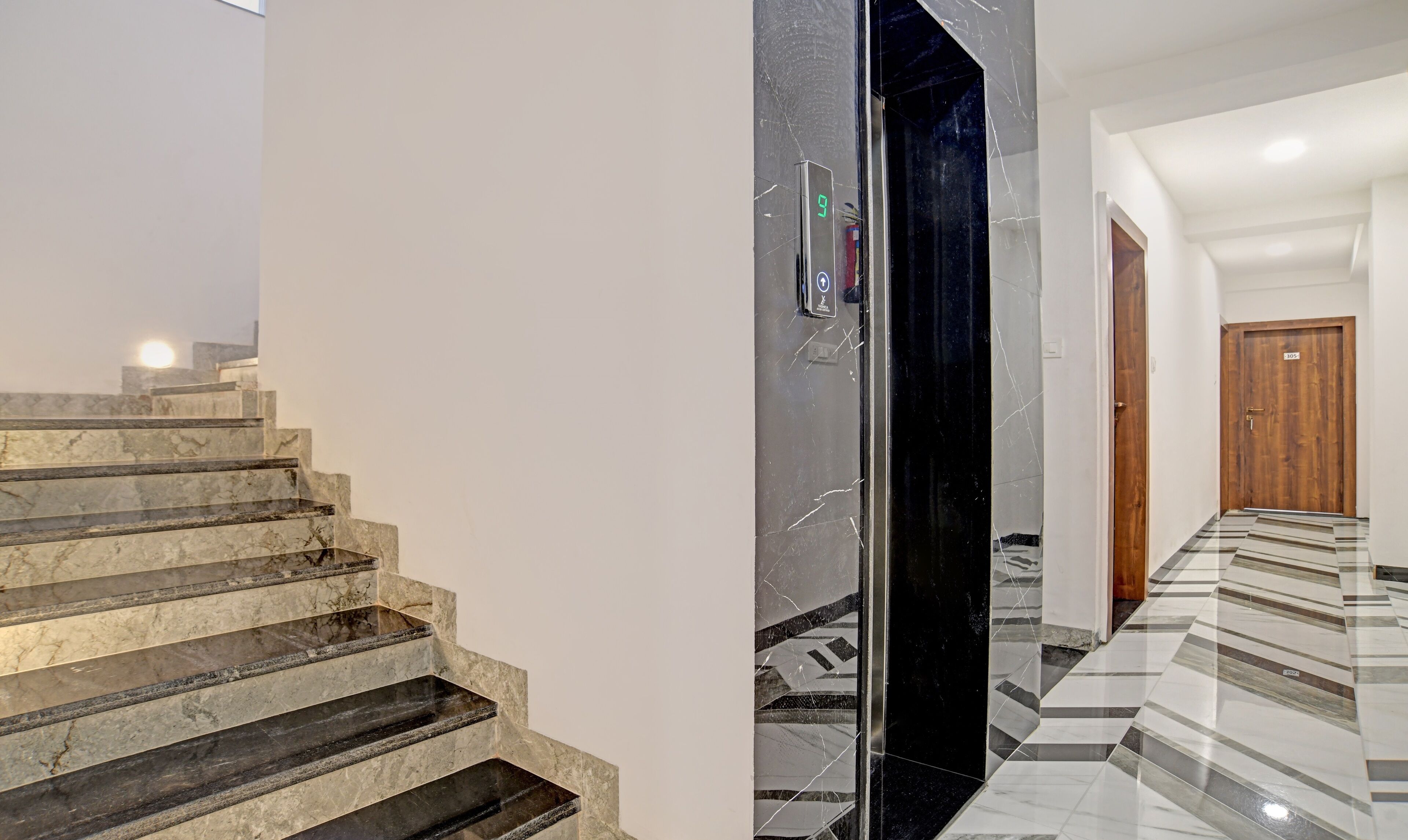A modern hotel interior with marble stairs, a black elevator, and a tiled hallway leading to a wooden door.