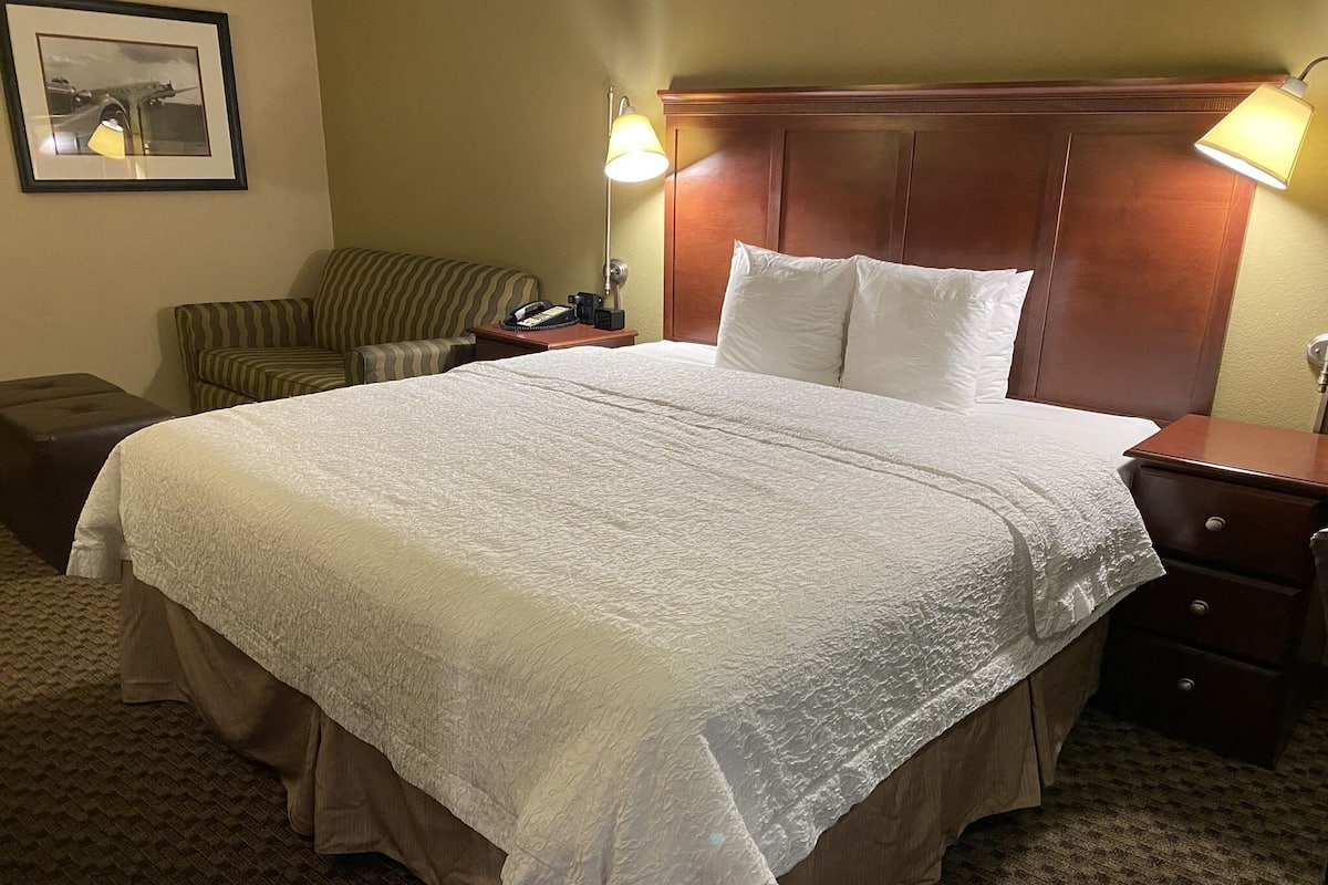 Une chambre d’hôtel avec un grand lit, une tête de lit en bois, une table de chevet avec une lampe, une chaise à rayures et un petit pouf.