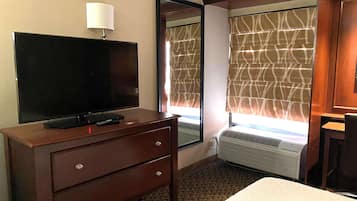 Habitación de hotel con televisor de pantalla plana sobre un tocador de madera, un espejo, una ventana con cortinas estampadas y una cama.