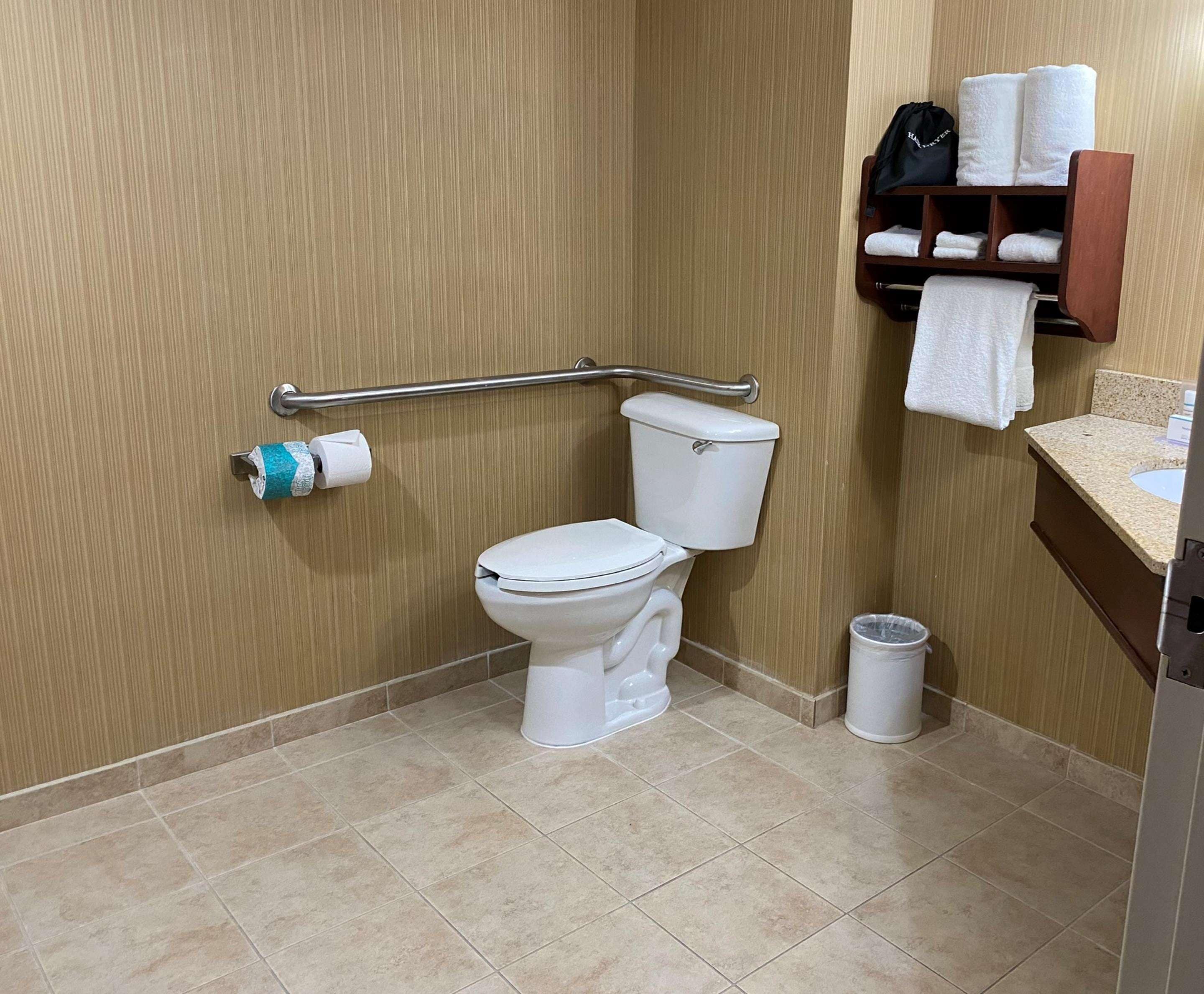 A bathroom with a white toilet, a grab bar, a towel rack with towels, and a trash can.