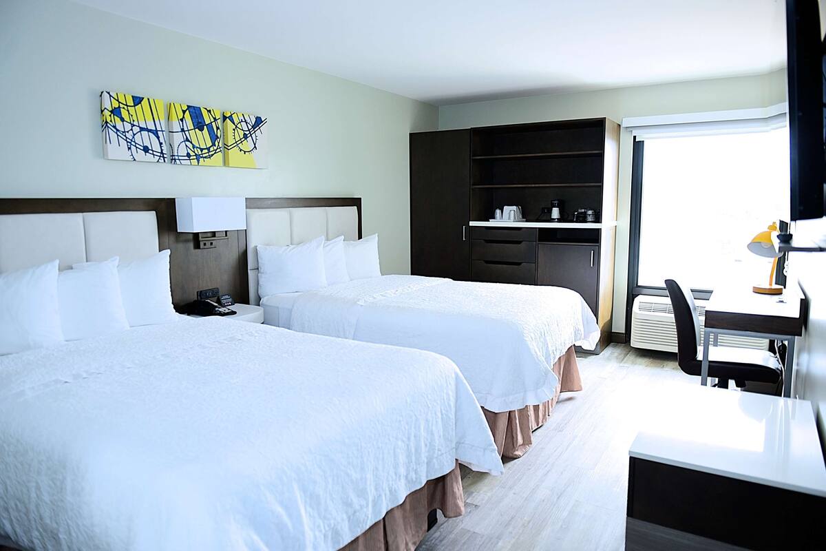 Une chambre d’hôtel avec deux lits, un bureau, une chaise, une armoire et un tableau au mur.