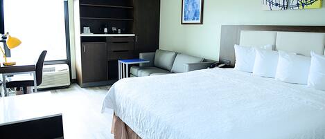 A hotel room with a large bed, a desk with a chair, a sofa, a wardrobe, and a painting on the wall.