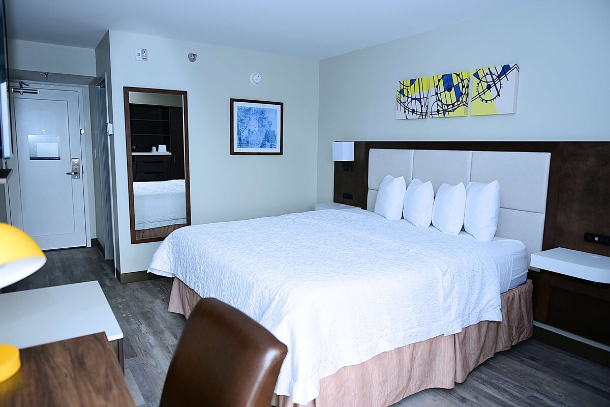 Ein Hotelzimmer mit einem großen Bett, einem Nachttisch, einem Sessel, einem Schreibtisch, einem Spiegel und Bildern an der Wand.