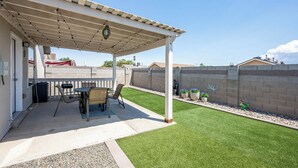 Een overdekt terras met een tafel en stoelen, een kleine tuin met kunstgras, en een bakstenen muur.