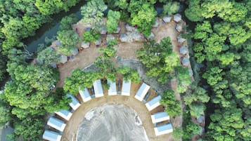 Luchtbeeld van een camping met tenten in een halve cirkel rond een open veld, omgeven door weelderige begroeiing en een rivier.