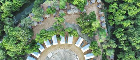 An aerial view of a campsite with tents arranged in a semi-circle around a central open area, surrounded by dense greenery and a river.