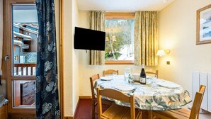 A dining area with a table set for two, a television mounted on the wall, and a window with curtains. - Apartment, les Avanchers Valmorel, sleeps 6. (Les Avanchers-Valmorel)