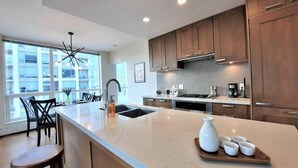 A modern kitchen with wooden cabinets, a large island, and a dining area with chairs. - Luxury Condo by River w/City Views (Calgary)