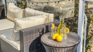 Un coin salon extérieur en osier, agrémenté de coussins, une table ronde avec une bouteille et des citrons, et des guirlandes lumineuses.
