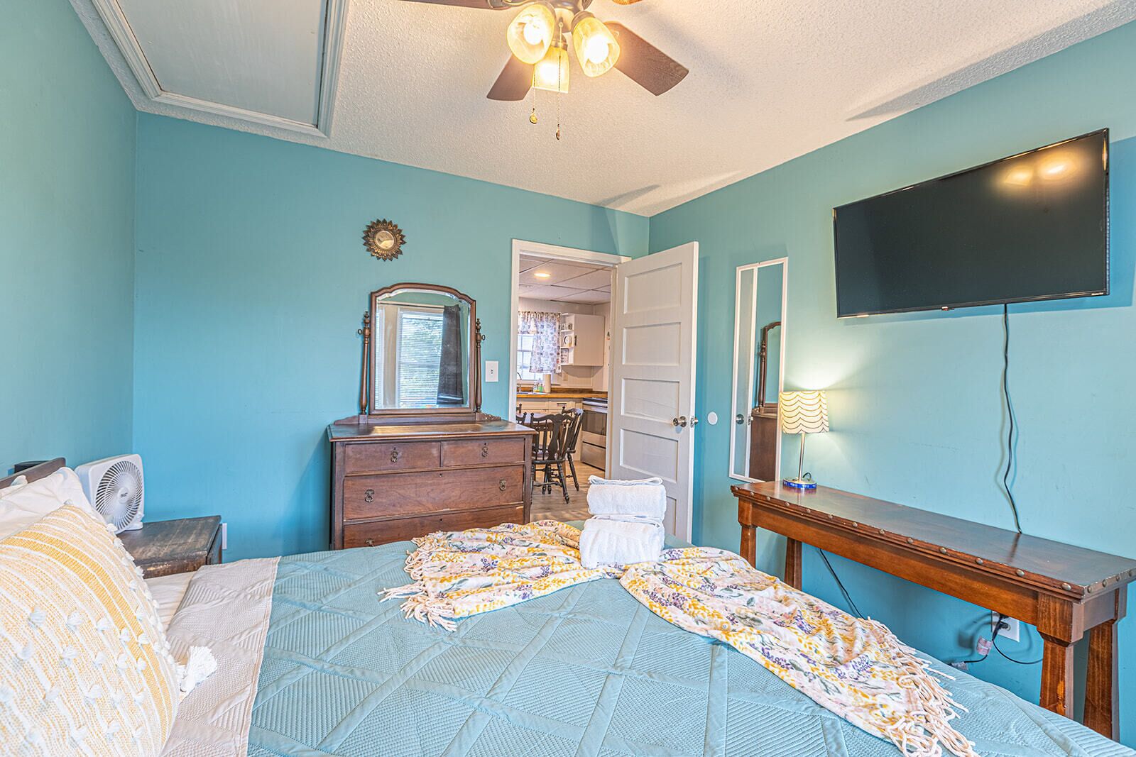 Une chambre à coucher comprenant un lit, une commode, un miroir, un ventilateur de plafond et un téléviseur fixé au mur.
