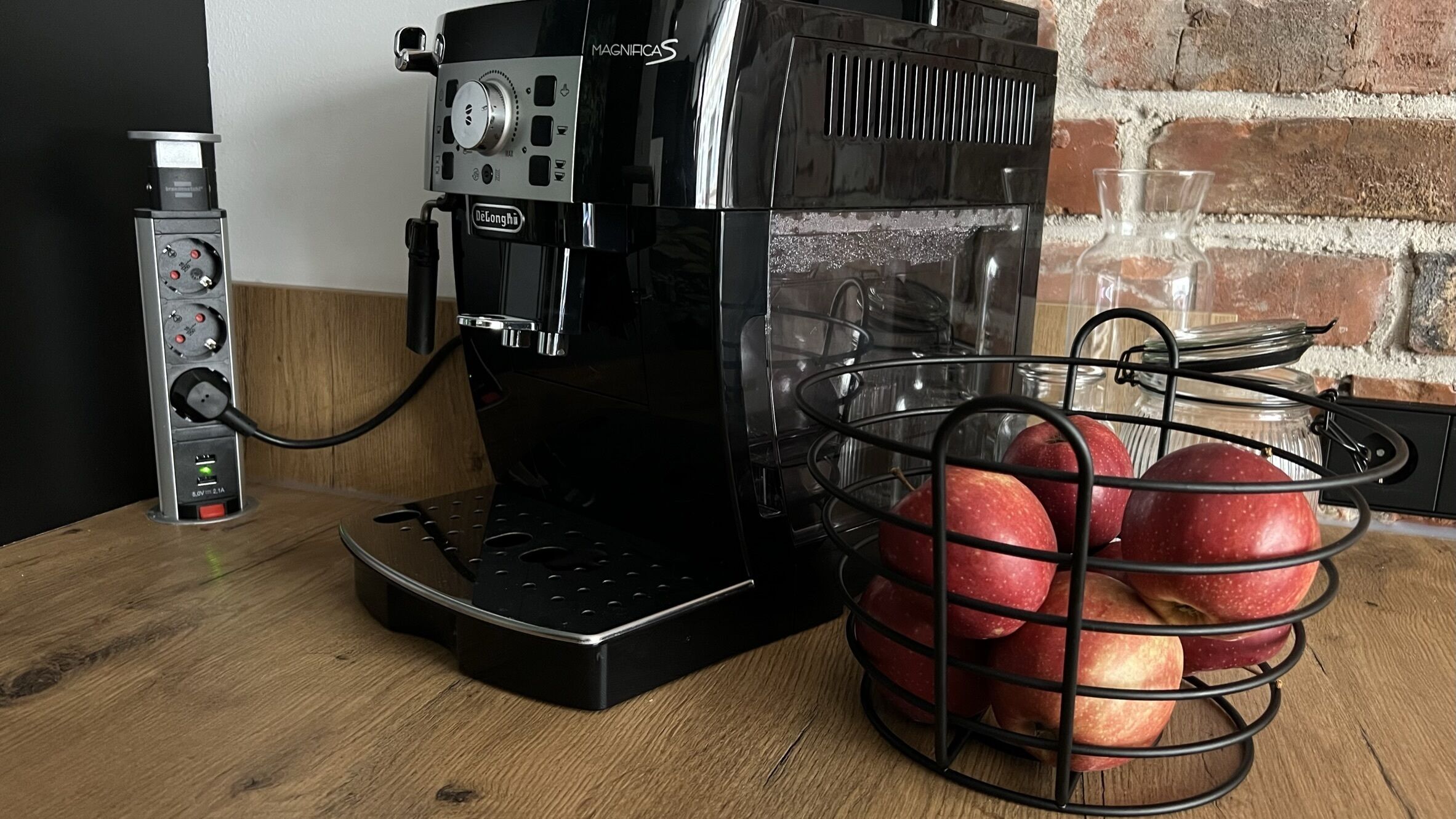 En kaffemaskine med en kurv æbler på et træbord.