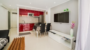 A modern living room with a TV, a white console table, and a dining area with a red kitchen in the background.
