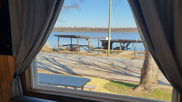 A view from a window with curtains, featuring a wooden bench, a table, and a dock extending into a body of water.