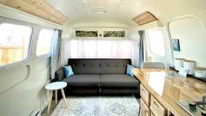 Een compact interieur van een camper met een slaapbank, een klein tafeltje, een houten dressoir en een raam met gordijnen.