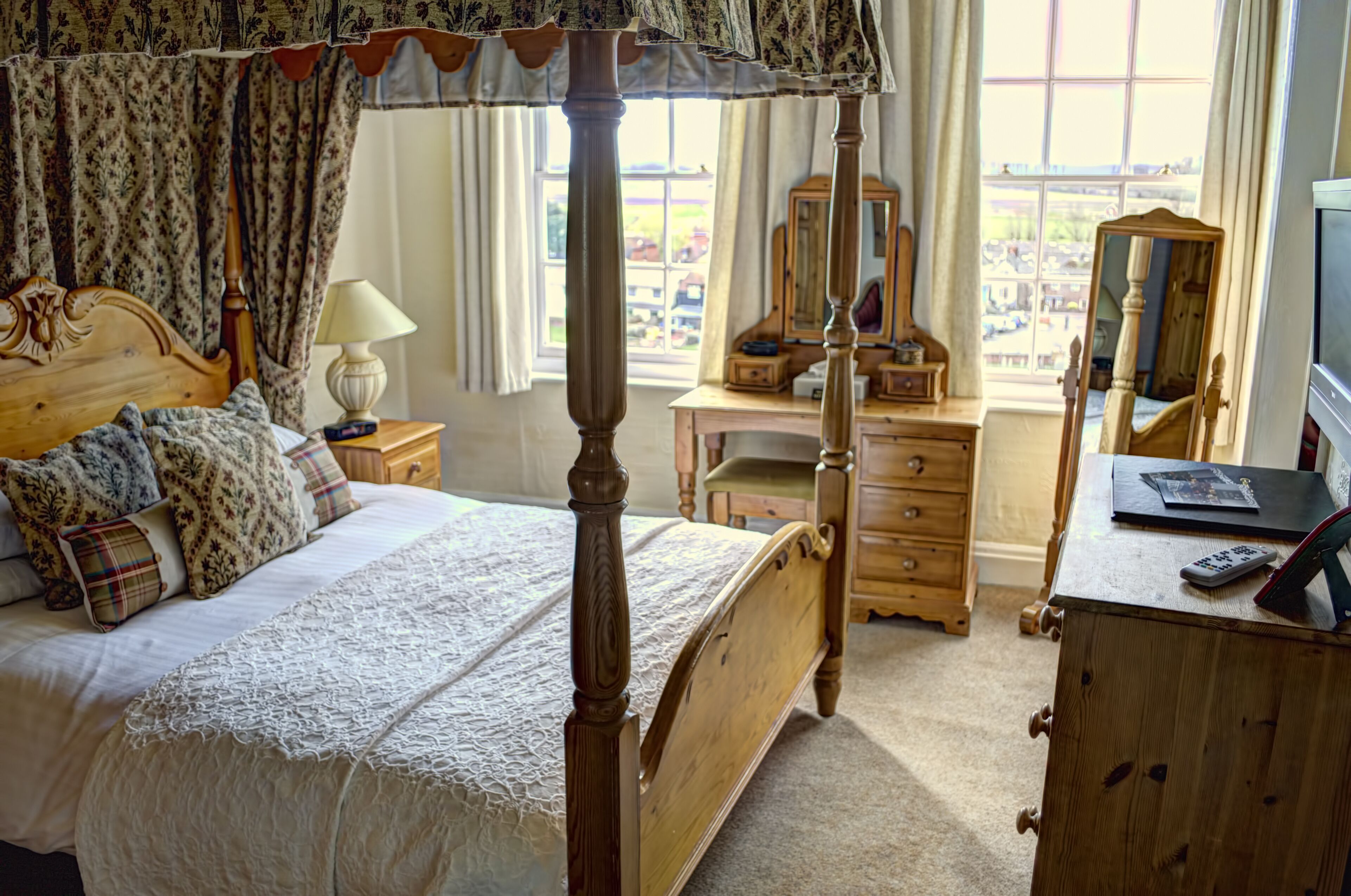 A traditional bedroom with a four-poster bed, a wooden dresser, a vanity with a mirror, and a desk with a laptop and remote control.