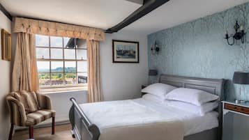 A bedroom with a bed, a chair, a nightstand, a lamp, a window with curtains, a painting, and a wall with a tree pattern.