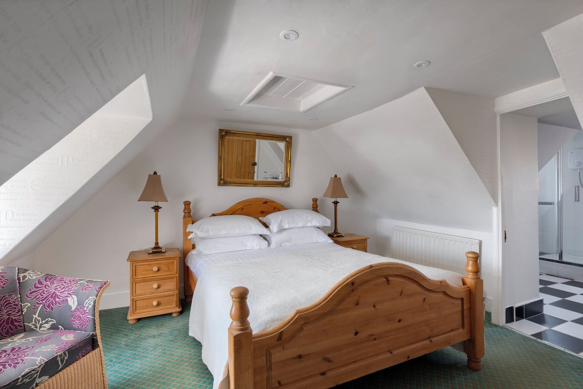Un dormitorio con techo inclinado, una cama de madera, dos mesitas de noche con lámparas, un espejo y un suelo a cuadros.