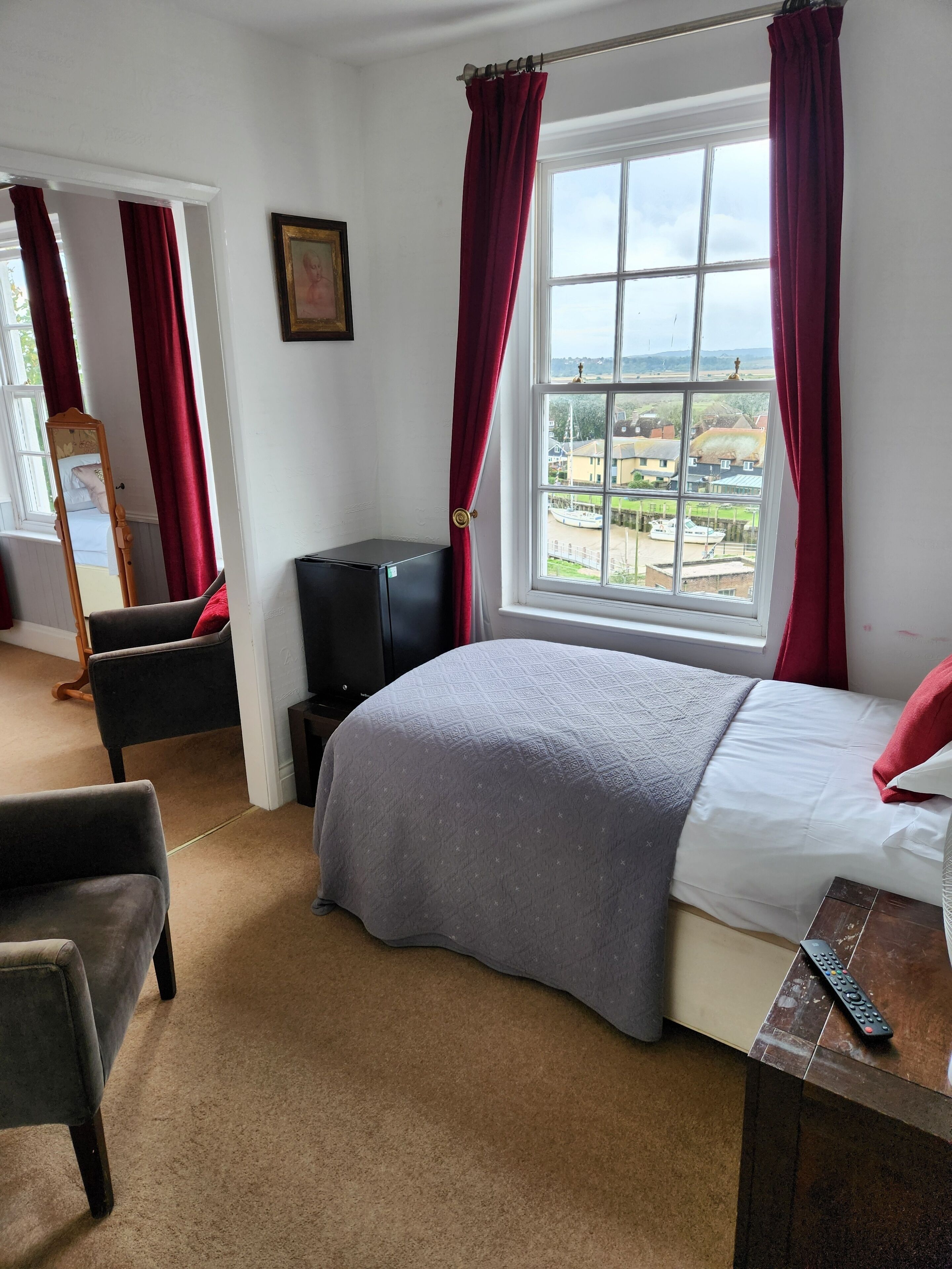 A bedroom with a bed, a chair, a desk, a mirror, and a window with a view of houses and boats.