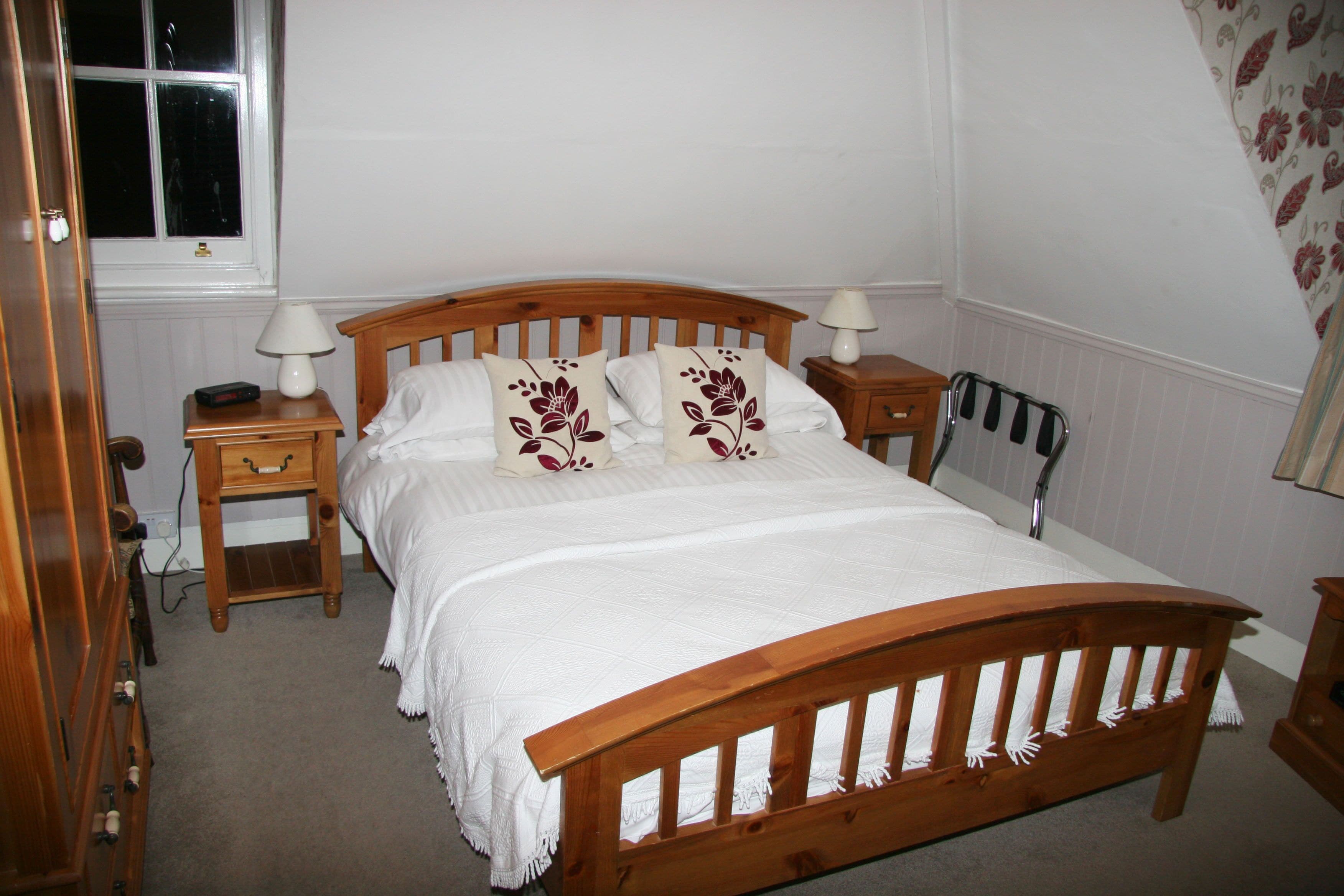 A neatly made bed with a wooden headboard, two bedside tables with lamps, a radiator, and a window with a view of the outside.