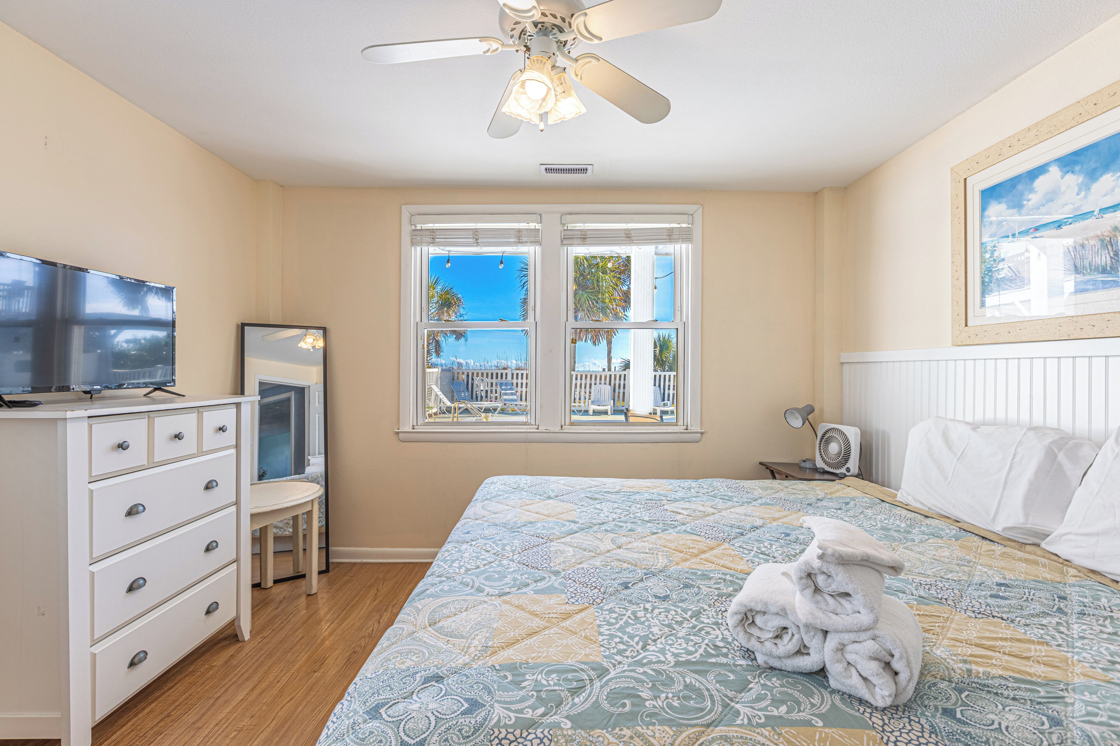 Un dormitorio con cama, ventilador de techo, televisor, cómoda y ventana con vistas a palmeras y una silla afuera.