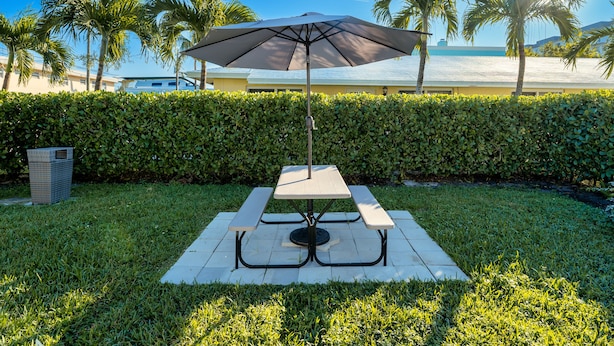 A picnic table with a large umbrella, surrounded by a well-maintained hedge, palm trees, and a building in the background. - PRAIA Hotel Hollywood (Hollywood)