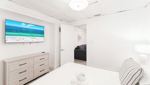 A hotel room with a large flat-screen TV on the wall, a bed with a white comforter, a wooden dresser, and a gray sofa in the background. - PRAIA Hotel Hollywood (Hollywood)