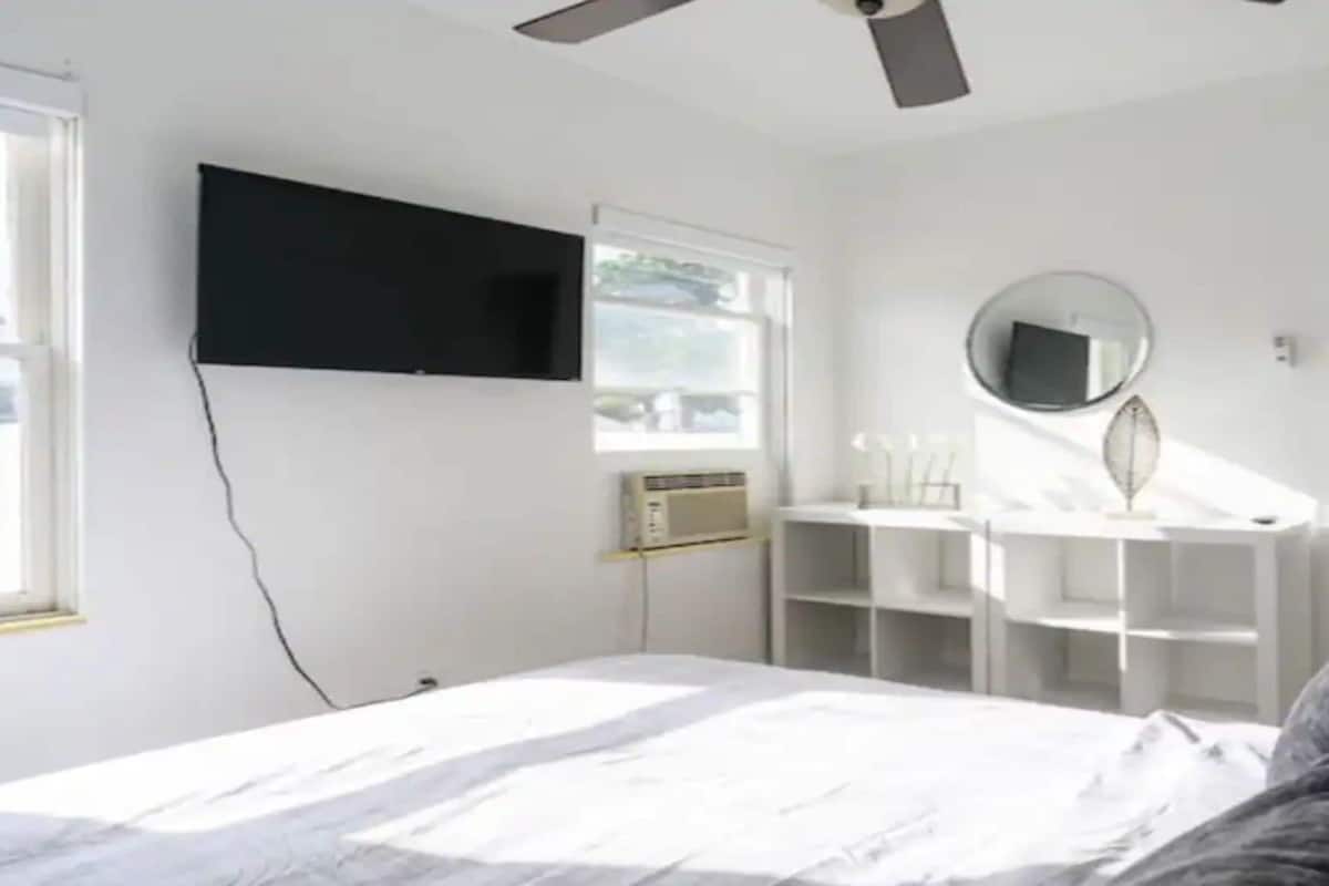 Une chambre à coucher avec un lit, un ventilateur de plafond, un téléviseur fixé au mur, un miroir et une porte.