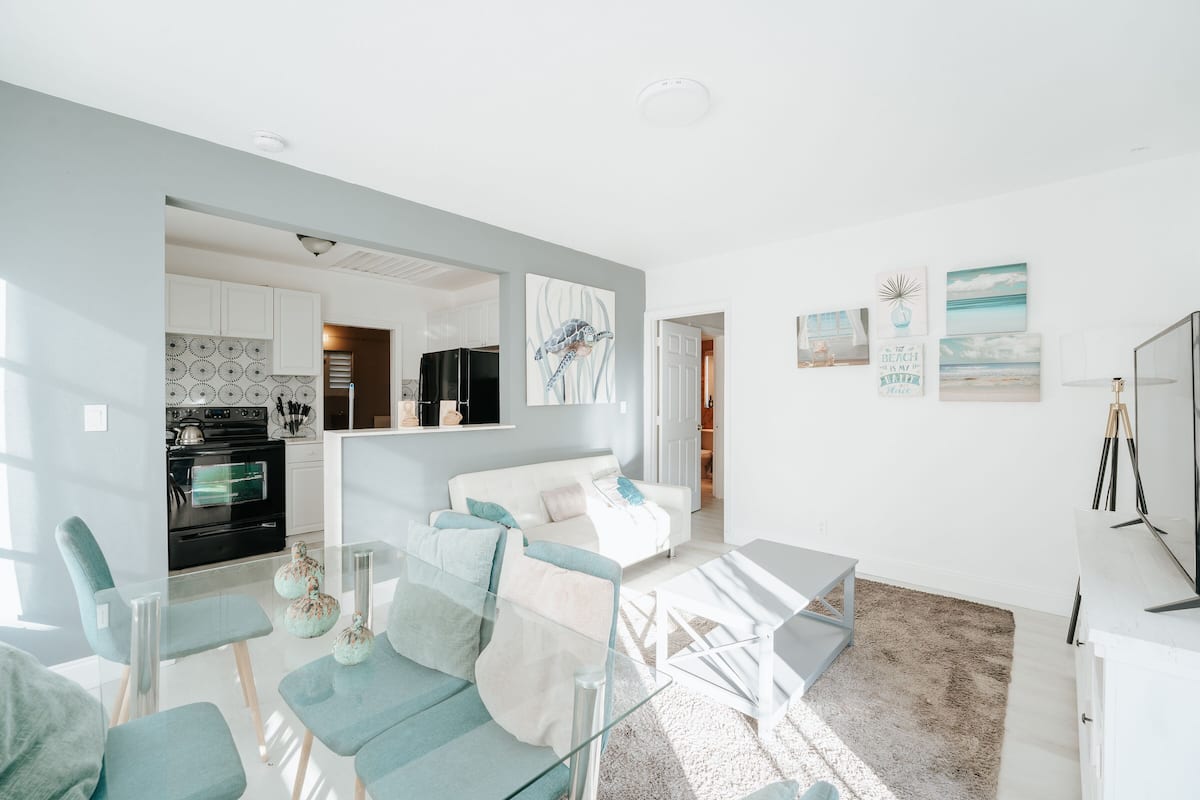 Un salon moderne avec une table à manger en verre, des chaises turquoise, un canapé blanc et une cuisine en arrière-plan.