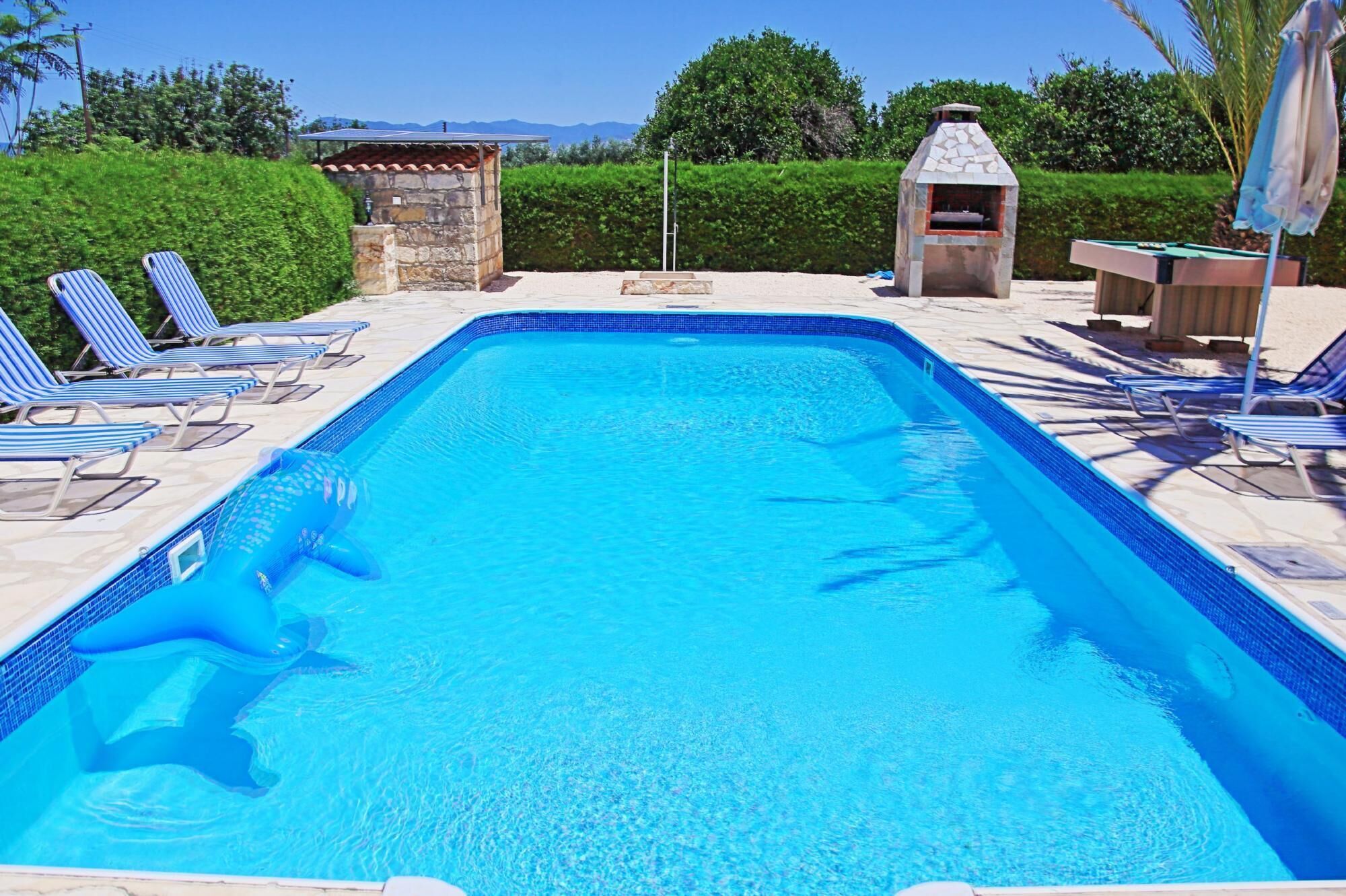 A rectangular swimming pool with clear blue water, surrounded by lounge chairs and a stone structure with a chimney.