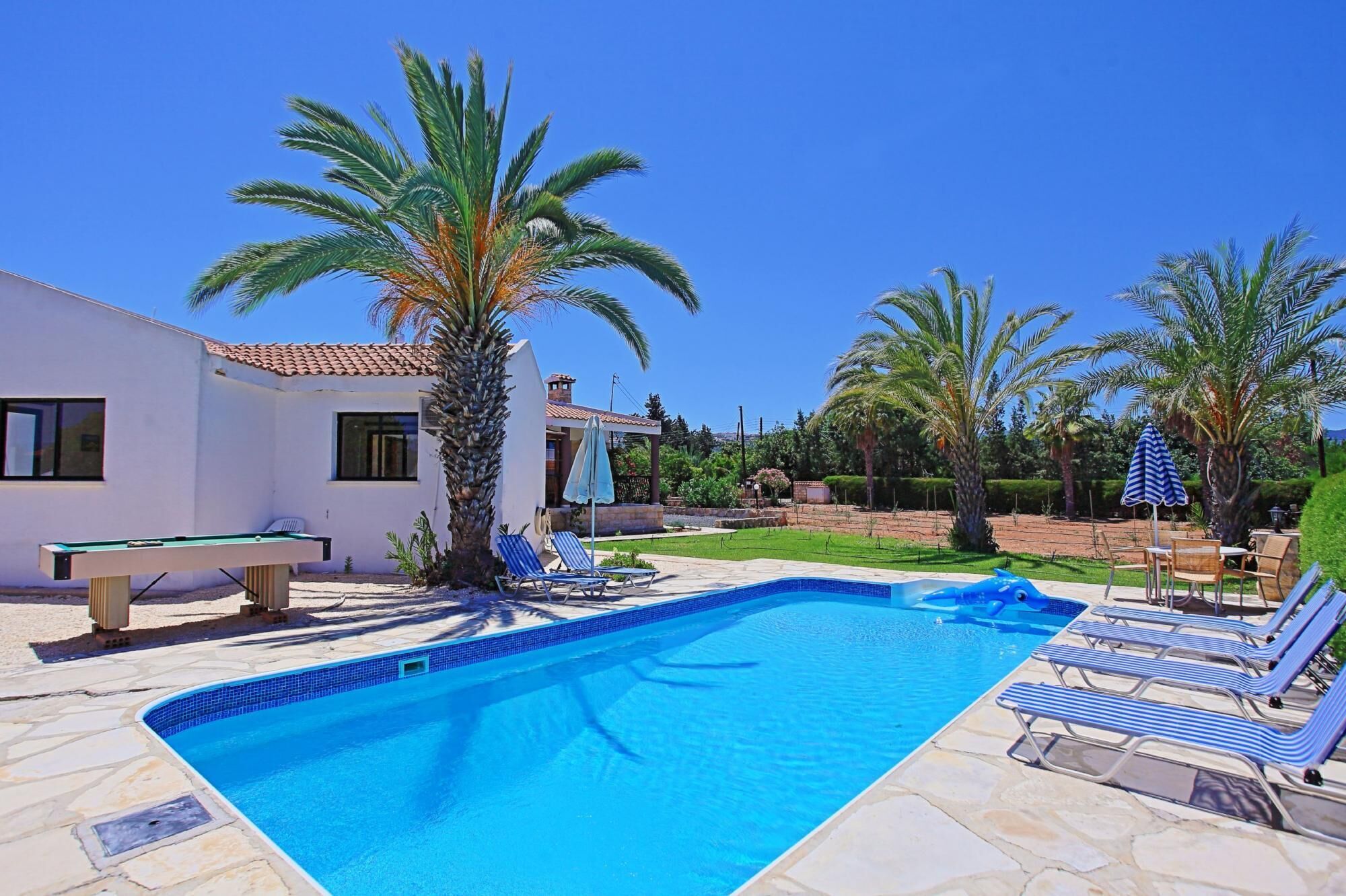 A swimming pool with lounge chairs and a pool table, surrounded by palm trees and a white building.