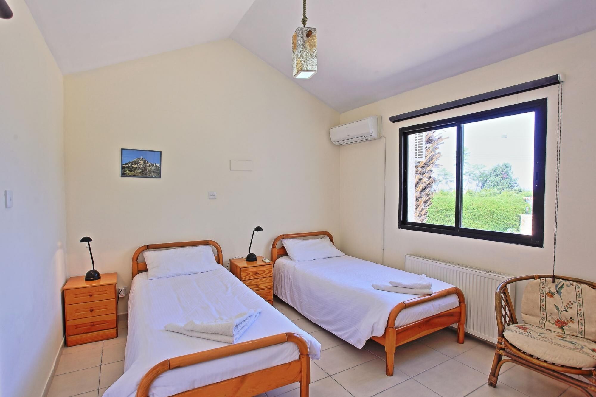 A room with two single beds, a wooden nightstand with a lamp, a chair, and a window with a view of greenery.
