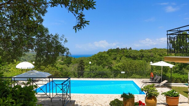 A swimming pool with a stone deck, surrounded by greenery and a view of the sea. - Villa Joanna in (Corfu)