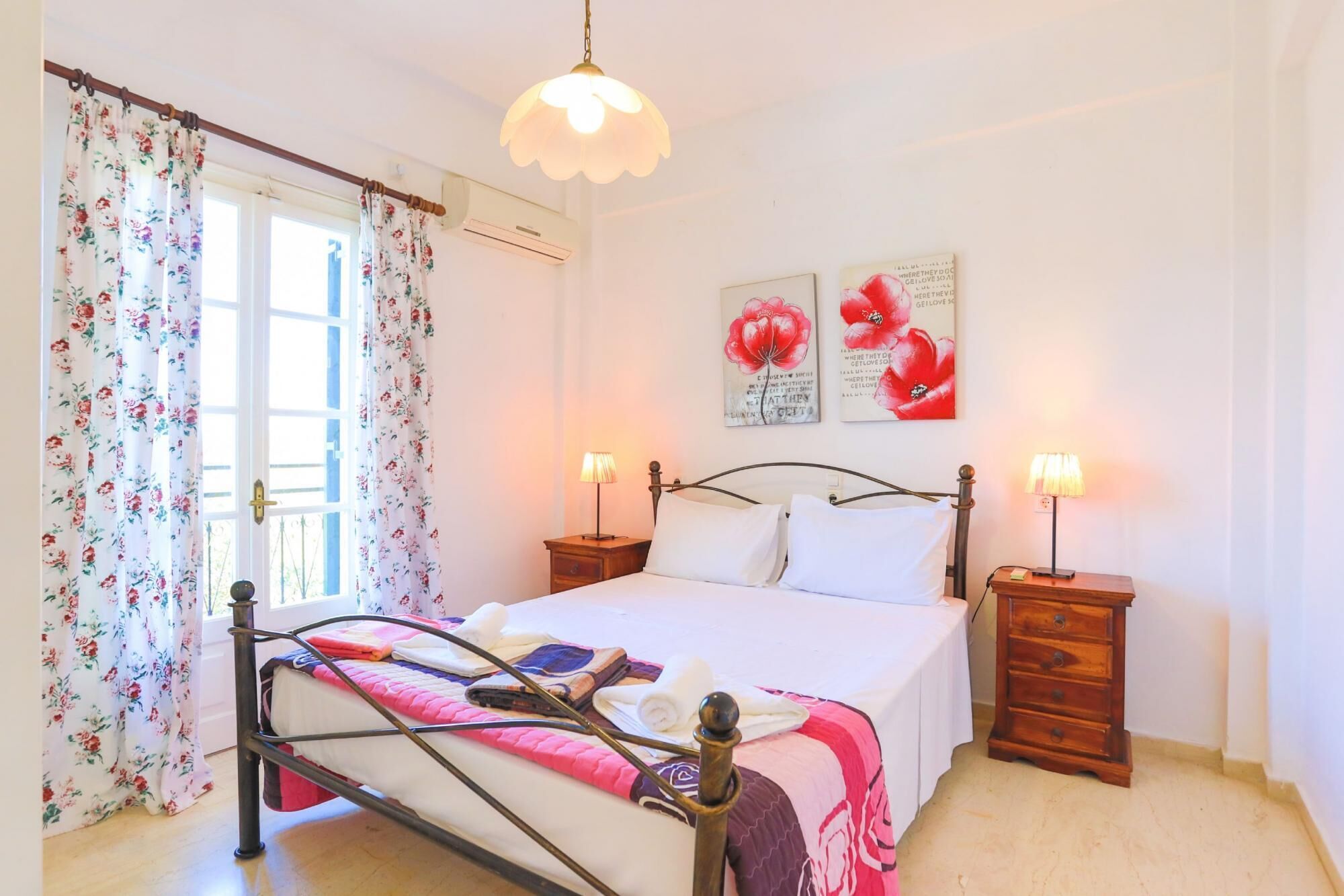 A bedroom with a bed, two bedside tables, a window with floral curtains, and two wall-mounted lamps.