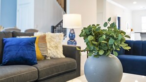 Un salon avec un canapé gris, un coussin bleu et un vase blanc orné de feuillage vert.