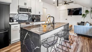 Una cucina moderna con mobili bianchi, un'isola in marmo bianco e nero, un fornello a gas e un forno a microonde integrato. Sono presenti sgabelli da bar e una TV montata a parete.