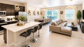 Eine moderne Küche mit dunklen Schränken, einer weißen Kücheninsel und Barhocker. Es gibt einen Essbereich mit einem beigefarbenen Sofa und einem Couchtisch.