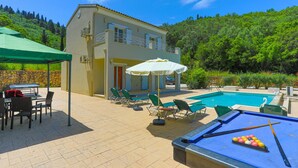 A pool area with a pool table, lounge chairs, and a two-story building with a stone wall. - Villa Anamari in (Corfu)