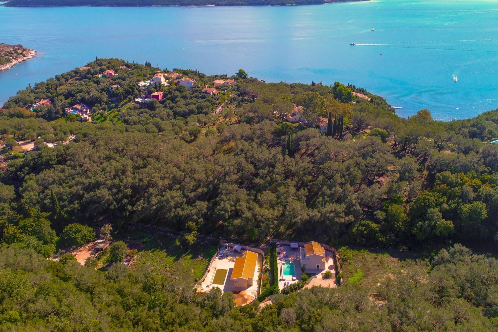 An aerial view of a coastal area with a cluster of houses surrounded by dense forest, a clear blue sea, and a lighthouse in the distance.