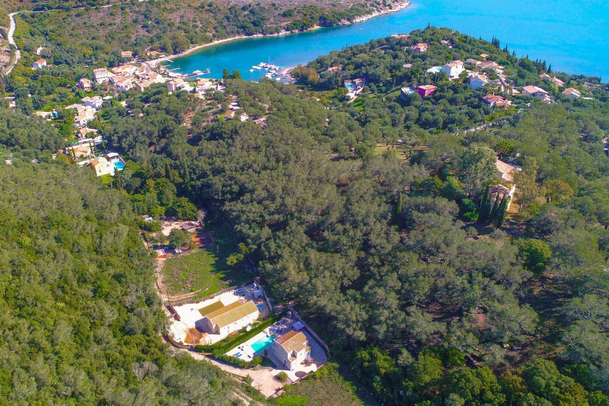 Vista aérea de uma área costeira com uma grande villa cercada por floresta densa, uma pequena praia e um porto com barcos.