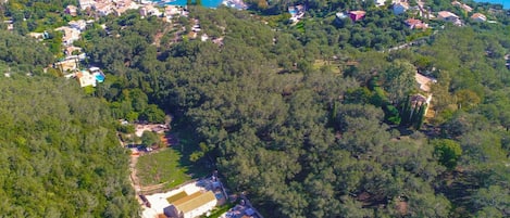 Vista aérea de uma área costeira com uma grande villa cercada por floresta densa, uma pequena praia e um porto com barcos.